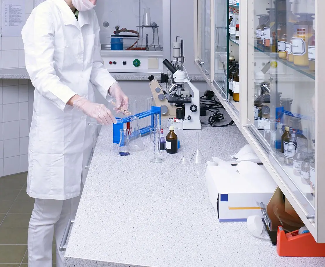 A lab technician wearing a white coat and gloves works in a laboratory, handling test tubes near a microscope and various chemical containers on a countertop.