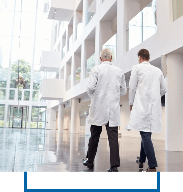 Two medical professionals in white lab coats walk together through a modern, spacious building with tall windows and glossy floors.