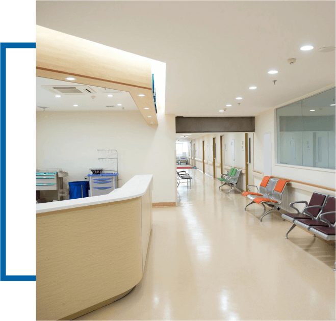 A hospital corridor with a waiting area, chairs lined up against the wall, a reception desk to the left, and bright overhead lighting.